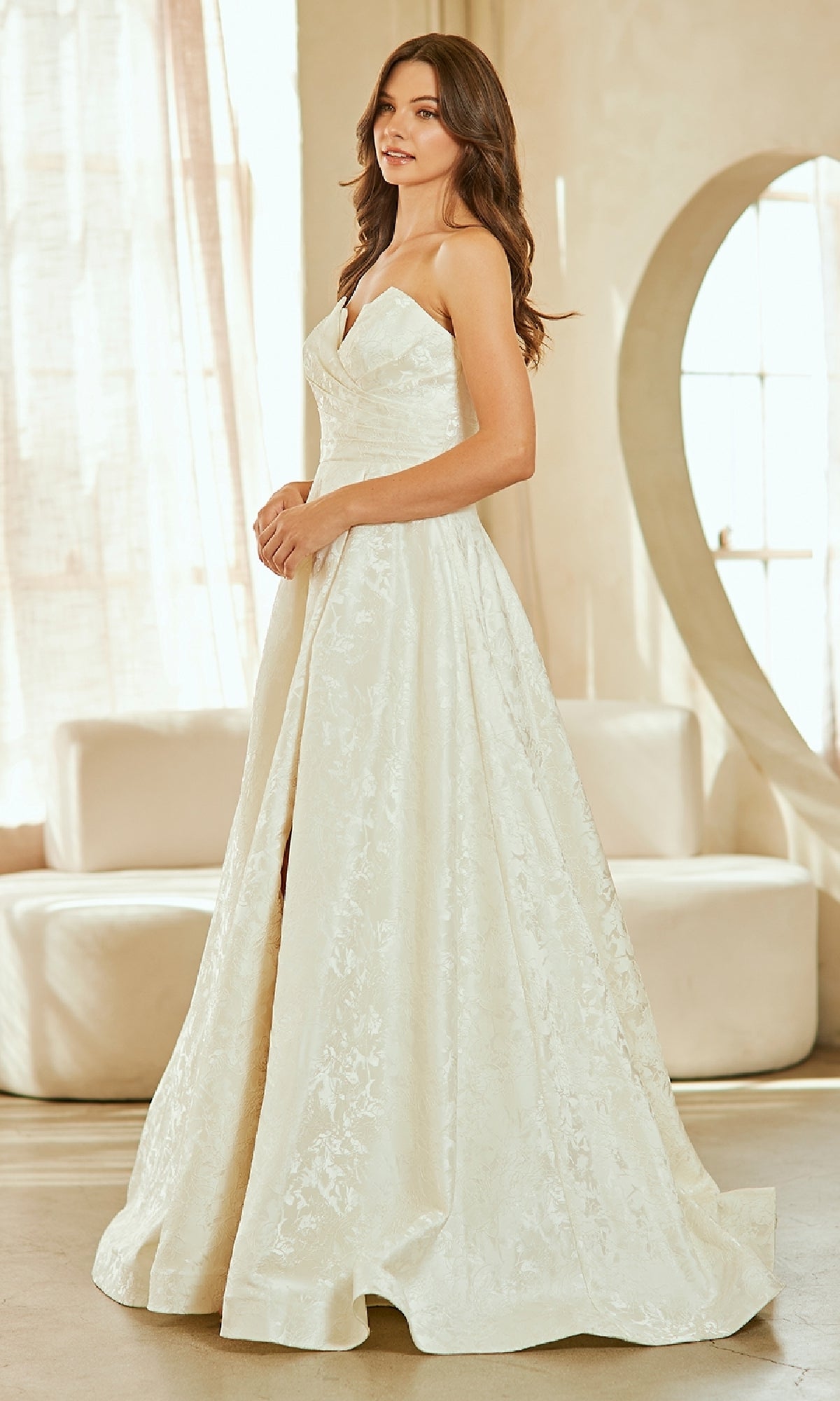 A woman in a textured strapless prom gown, Amelia Couture 8061, stands indoors by a large window and modern white furniture.